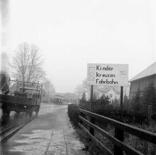 Hauptstraße: rechts Zaun. dahinter in Vorgarten Hinweisschild auf Kinder: links Lkw: hinten Mitte parkender Lkw und Pkws: hinten rechts und links Wohn- und Geschäftshäuser