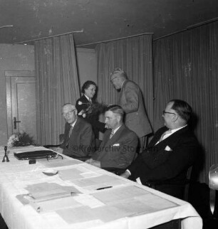 Keck, Erika: Kreispräsidentin: Kreisberufschule Bad Oldesloe: Kreistagssitzung am 18. Mai 1955: Verabschiedung und Überreichung eines Blumenstraußes durch Landrat Wilhelm Siegel: Präsidiumstisch mit dem neuen Kreispräsidenten Friedrich Hardt und den Stellvertretern Willy Esser und Paul Manthey: hinten Vorhänge