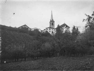 In Kaltern : Blick über Apflebäume und Weinhänge auf die Pfarrkirche Maria Himmelfahrt in Kaltern