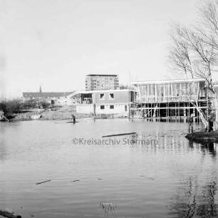 Am Exerzierplatz: vorne der Teich mit Person auf dem Wasser, rechts dahinter das im Bau befindliche Sportlerheim, links davon das Hausmeisterwohnheim, im Hintergrund das Rathausgebäude und Wohngebäude der Greifswälder Kehre: Blick nach Norden