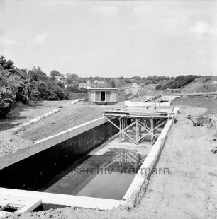 Zentralklärwerk: Anna-Heitmann-Weg: Belebungsbecken und Pumpwerk nach Fertigstellung: Baugrube zugeschüttet, links offener Betonkanal von Pumpenhäuschen hinab: Becken mit Isolieranstrich, in vorderem Becken Gerüst mit Arbeitsbühne, hinten Arbeiter auf Beckenrand: Baumaterial: hinten Trave, an gegenüberliegendem Ufer Häuser