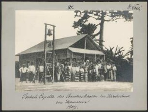 Gottesdienst in Protestantischer Kapelle, Kamerun : Portrait einer großen Menschengruppe vor einem Gebäude, im Vordergrund ein externer Kirchturm als Holzkonstruktion, fotografische Reproduktion nach gemalter Vorlage