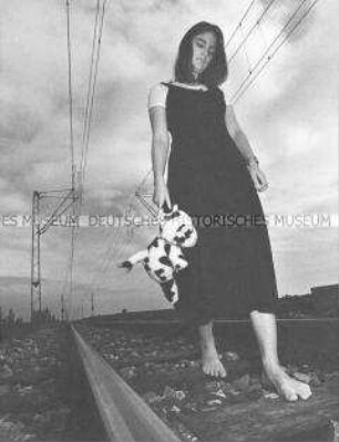 Mädchen mit Kuscheltier an der Hand läuft barfuß an Bahnschienen entlang (Altersgruppe 18-21)