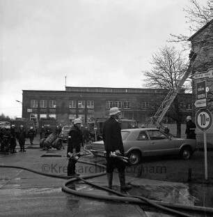 Brand: Gies Kerzen GmbH: Möllner Landstraße: Feuer in Parafinturm: vorn zwei Feuerwehrmänner mit Kupplungen zwischen Wasserschläuchen: rechts dahinter Pkw und Löschfahrzeug mit Leiter: darauf Feuerwehrmann: hinten weitere Löschfahrzeuge und Feuerwehrleute: im Hintergrund Gewerbehalle