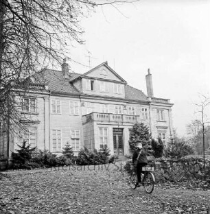 Grönwohldhof: Gutshaus: vorn Auffahrt: darauf Postbotin mit Fahrrad: rechts Mauer aus Feldsteinen: links Baum und Tannenreihe