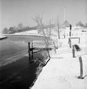 Trave: zugefrorener Fluss: in der Mitte Bootsanleger hinter Bäumen: rechts Zaun und Fahnenmast: dahinter eingezäunte Weiden: hinten rechts Haus: im Hintergrund Bäume: Winter