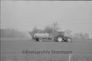 Landwirtschaft: Traktor mit Güllewagen auf Feld: im Hintergrund Stromleitungen und Bäume