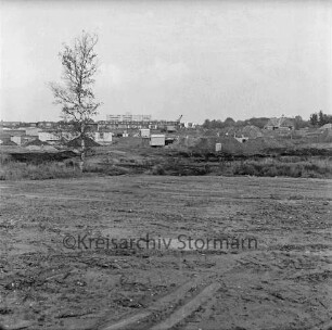 Dammannsche Koppel: Neubaugebiet: vorn planiertes, dahinter unbefestigtes Gelände mit Erdhaufen, Bauwagen, Fundamenten und Rohbauten von Wohnhäusern: dahinter Reihenhäuser und Wohnblock: 15. Oktober 1976