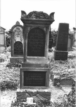Grabstein 62 (Index: 99808311) Material: Sandstein, schwarze Marmorplatte Zustand: leicht beschädigt Maße HxBxT: 179x62x33 cm Beschreibung: Stele, getreppter Sockel, unterer Sockelteil rusticaartig, Aufsatz mit Voluten und Eckakroteren, Schriftplatte Symbolik: Blätter, Eicheln: Grabstein 62