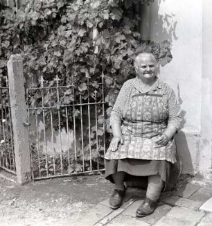 Landleben. Oma sitzt : Wasewitz. Eine alte Frau sitzt auf einem Stuhl vor einem Haus, dass mit Wein berangt ist