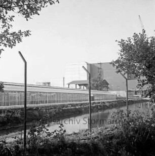 Gewächshäuser: dahinter der Gebäudekomplex der Müllverbrennungsanlage: vorn Wasserbecken, Pfähle eines Sicherheitszaunes sowie Sträucher und Bäume
