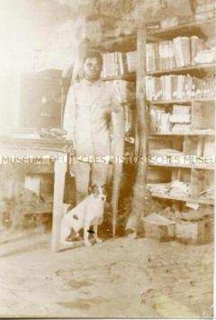 Afrikanischer Mann in europäischer Kleidung posiert mit Elefantenstoßzähnen und einem Hund vor einem Bücherregal