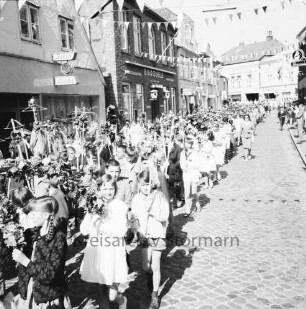 Kindervogelschießen: Umzug: Hindenburgstraße: Mädchengruppen mit Blumenbögen, Jungen mit Blumenstäben in Armbrustform und Fähnchen: an den Straßenrändern Zuschauer: über Straße Wimpelgirlanden: an linker Straßenseite Friseursalon Lübcke, Gaststätte "Bierquelle": hinten Hagenstraße, Toreinfahrt zu Holz- und Baustoffhandlung Hugo Eggers, Gasthof "Stadt-Krug", 28. Juni 1965
