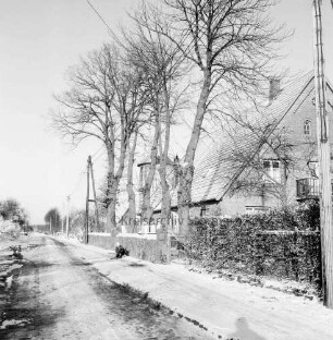 Dorfstraße mit Kopfsteinpflaster: rechts Bauerngehöft mit Silo: davor Vorgarten und Baumreihe: vorn Kind mit Schlitten: Winter