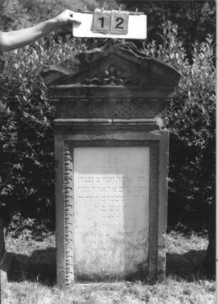Grabstein 12 (Index: 100113793) Zustand: leichte Beschädigungen am Giebel und am Rand Maße HxBxT: 138x65x30 cm Beschreibung: Stele mit Spitzgiebel, Eierstabfreis um Textteil, Kettenfries am Giebel, vertiefter Textteil Symbolik: 3 Mohn(?)blüten mit Band im Giebel: Grabstein 12