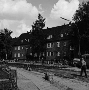 Hebbelstraße: Ausbau: vorn unbefestigte Straße, Absperrung, Stapel mit Gehwegplatten, Mülleimer, zwei Arbeiter und Heck eines Kleinbusses mit Anhänger: dahinter Wohnhäuser der Gemeinnützigen Baugenossenschaft Bad Oldesloe mit Vorgärten