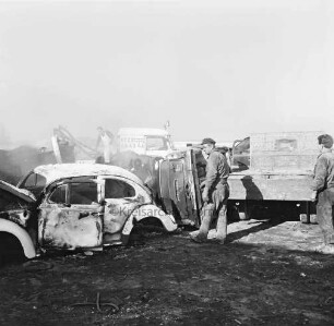 Harry Kloss Autoverwertung GmbH & Co. KG: Kampstraße: Abstellplatz mit Schrottautos: vorn rechts zwei Arbeiter vor Lkw: links VW-Käfer: dahinter Mann auf Fahrzeug eines Bergungsdienstes beim Abladen eines Pkws