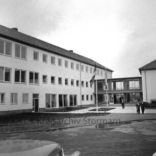 Ahrensburg: Hermann-Löns-Straße: Erweiterungsbau: Einweihung: Außenansicht: Gäste auf Vorplatz: Fahnenmasten: hinten rechts Verbindungstrakt mit Eingangsportal, Altbau, 26. Juni 1962
