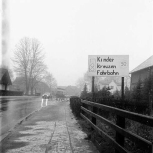 Hauptstraße: rechts Zaun. dahinter in Vorgarten Hinweisschild auf Kinder: hinten Mitte Pkw: dahinter parkende Lkw und Pkws: hinten rechts und links Wohn- und Geschäftshäuser