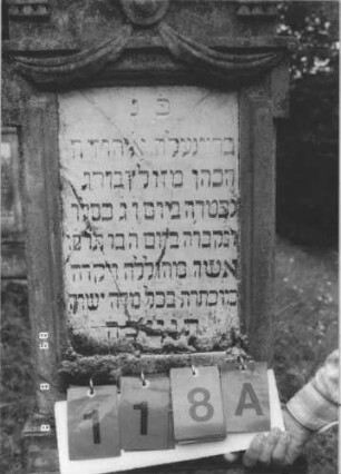 Grabstein 118 (Index: 99800082) Material: Sandstein, Schriftplatte Marmor Zustand: verwittert, Schriftplatte zersprungen Maße HxBxT: 154x46x21 cm Beschreibung: Stele, Giebel mit Verdachung, rechteckige eingelassene Schriftplatte, getreppter Sockel: Grabstein 118A