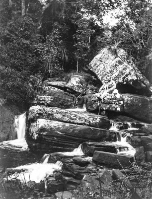 Wasserfall : Blick auf einen kleinen Wasserfall bei Bambuli. Auf einem dieser Felsen ein Mitglied der Expedition in Tropenkleidung mit einem Hund auf dem Schoß.