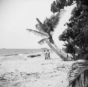 Strandpanorama : Zwei Männer gehen den Strand bei Bata entlang, im Hintergrund auf dem Ozean Frachter.