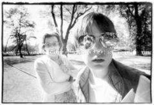 Szene in einer Parkanlage: Junges Mädchen mit Pilotenbrille, in der sich der Fotograf spiegelt, hinter ihr eine lachende Dame (Altersgruppe 18-21)
