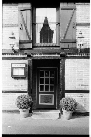 Restaurant Zum Rauchhaus Anno 1756: Bei der Alten Kate: Außenansicht: Fachwerkhaus [ehemalige Ahlemann'sche Kate]: Giebelseite: Detailaufnahme: Eingangstür, daneben Kasten mit Speisekarte, beiderseits Laternen, darüber Fenstertür mit Geländer und Holzläden