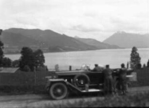 Deutschland? Blick über einen See gegen Gebirge. Im Vordergrund Ausflügler neben einem Personenkraftwagen mit offenem Verdeck