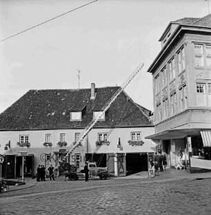 Hude: Feuerwehrgerätehaus "Blaues Haus" mit geöffneten Toren: in der Mitte Feuerwehrfahrzeug mit ausgefahrener Drehleiter: daran Feuerwehrmänner: rechts Wohn- und Geschäftshaus Gebrüder Petersen und Zuschauer