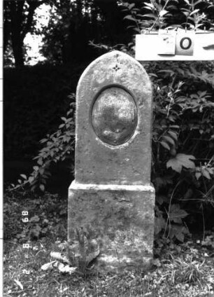 Grabstein 10 (Index: 100101722) Material: Sandstein, schwarze Marmorplatte Zustand: Schriftplatte verwittert mit starkem Textverlust Maße HxBxT: 108x35x12 cm Beschreibung: Stele, Sockel, Spitzgiebelabschluß, ovale Schriftplatte Symbolik: Sternchen: Grabstein 10