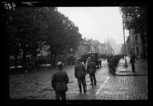 Tournai (Belgien): Personenkraftwagen mit geschlossenem Verdeck auf einer breiten gepflasterten Straße neben einer Baumreihe, am Straßenrand ein lockeres Spalier von Soldaten mit Stahlhelm, im Hintergrund die Silhouette der Kathedrale
