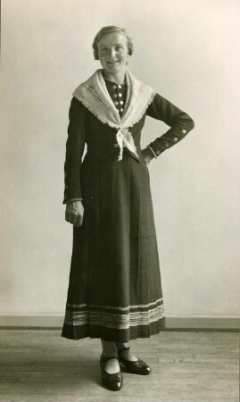Frau stehend, mit in der Taille gestützem linken Arm in Volkstracht mit handgewebtem Kleid. Frontal. Halbtotale