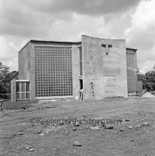 Katholische Heilig-Geist-Kirche: Wöhrendamm: Kirchengebäude vor Fertigstellung: Außenansicht von Südosten: Eingangsbereich mit Reliefs: hinten Mitte in Eingang Maler: vorn unbefestigte Außenanlage: hinten rechts und links Bäume
