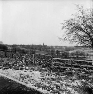 Travetal: links Gatter: dahinter Baum: hinten Trave: Blick über Wiesen und Knicks: im Hintergrund evangelische Kirche und Häuser: Winter