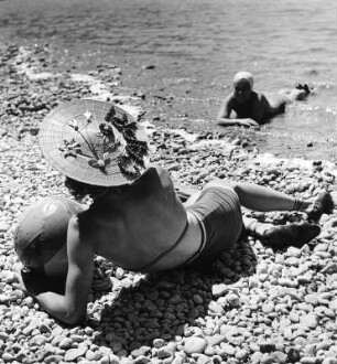 Badebilder. Frau mit Ball und Hut, im Badeanzug an einem steinigen Uferrand liegend