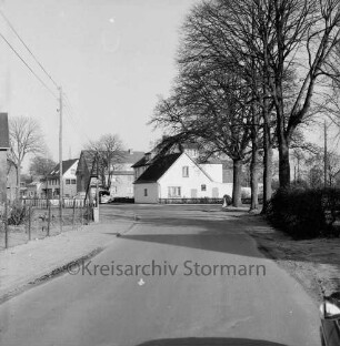 Hauptstraße: rechts Baumreihe hinter Zaun und Hecke: hinten Wohn- und Geschäftshäuser: davor parkende Pkws: links Zaun und Strommast: dahinter Garten