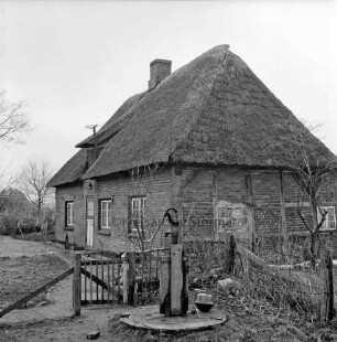 Armenhaus: kleines Haus mit Reetdach und Fachwerk: vorn Hecke mit Gatter: davor Pumpe