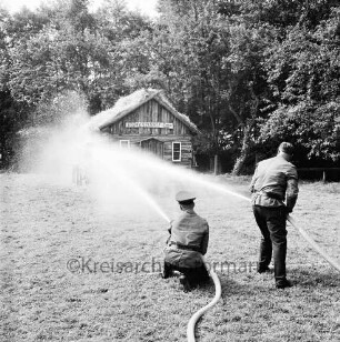 Kupfermühle: Amtsfeuerwehrfest: Löschen der "Kupfermühle Anno 1500": vorn zwei Feuerwehrmännder mit Wasserschläuchen: hinten brennendes Holzhäuschen: dahinter Bäume
