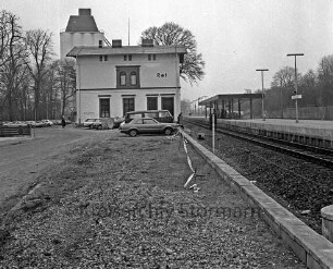 Bahnhofsgebäude, parkende Autos: rechts Bahnsteig, Gleise: im Hintergrund links Silo der Firma Ströh: im Vordergrund Freifläche mit Schotter, Absperrband