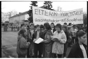 Emil-Nolde-Schule [Grundschule]: Am Schulzentrum: Elternprotest gegen Umwandlung in Gesamtschule: Marga Möller, Brunhilde Beeken und weitere Eltern beim Verteilen von Flugblättern, Schüler, Transparent "Eltern wacht auf. Gesamtschule ja - auf diese Art nein": hinten Schulhof, Eingangstor, Schulgebäude: 20. März 1990