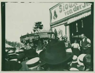 Zuschauer vor einer Schaubude mit "Sioux-Indianern" mit Karussell und Riesenrad