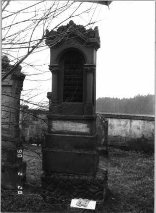 Grabstein 54 (Index: 100114898) Material: Sandstein und polierter Granit Zustand: Stele verrutscht, Texttafel mehrfach zerbrochen und repariert Maße HxBxT: 263x71x36 cm Beschreibung: Ädikula mit gestuftem Sockel auf Rustikasockel, eingezogener Spitzgiebel mit Akroteren und Laubdekor, eingesetzte Texttafel: Grabstein 54