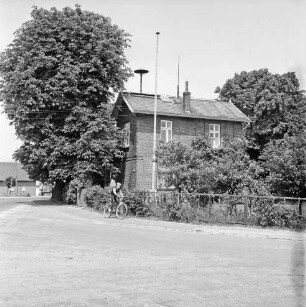 Dorfring: Wohnhaus mit Sirene auf dem Dach: ehemalige Volksschule: davor Vorgarten mit Hecke und Zaun: links davor Kastanienbäume: rechts Junge auf Fahrrad: links Landesstraße Richtung Autobahn A 1 und Stapelfeld: dahinter Bauernhaus
