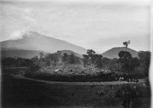 Siedlung : Blick von einer Anhöhe auf eine von Palisaden geschützte Siedlung.Im Hintergrund der Mount Meru von Wolken verhangen