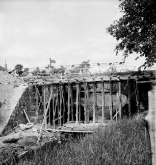 Heilsau: Brückenbau: Holzgerüste: darüber Absperrungen: vorn rechts und links Böschung, Büsche und Bäume