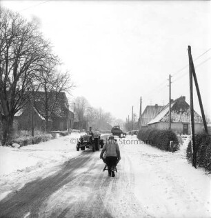 Dorfstraße mit landwirtschaftlichen Fuhrwerken: vorn Mann mit Schubkarre: hinten links Bäume: im Hintergrund Bauernhäuser: Winter