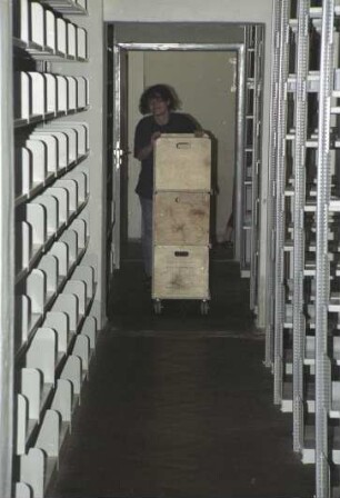 Sächsische Landesbibliothek - Staats- und Universitätsbibliothek Dresden. Zentralbibliothek (ehem. Landesbibliothek). Umzugsvorbereitungen im Magazin-Flachbau (Erdgeschoss). Hilfskraft mit Bücherwagen zwischen Regalen : Dresden-Neustadt, Marienallee 12. Sächsische Landesbibliothek - Staats- und Universitätsbibliothek Dresden. Zentralbibliothek (ehem. Landesbibliothek). Umzugsvorbereitungen im Magazin-Flachbau (Erdgeschoss). Hilfskraft mit Bücherwagen zwischen Regalen