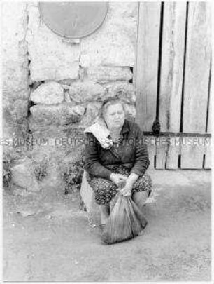 Eine Frau sitzt auf einem Stein, sie hält einen Einkaufsbeutel vor ihren Füßen, dahinter Steinmauer