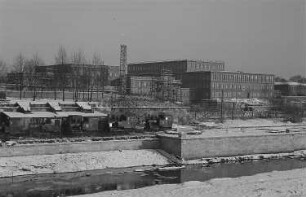 Industrieanlage im Winter, im Vordergrund ein Wasserlauf mit Befestigung und Werksbahnanlagen
